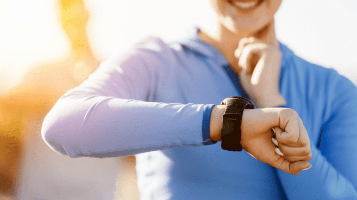 A person wearing a light blue jacket checks their smartwatch, appearing to take their pulse with the other hand. The sunlit, slightly blurred background suggests an outdoor setting where they treat themselves kindly, evident in their warm smile.