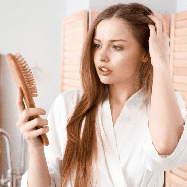 A woman with long brown hair in a white robe looks concerned as she holds a hairbrush filled with strands. Standing in her bathroom before wooden shutters, shes contemplating the best shampoos for women with thinning hair.