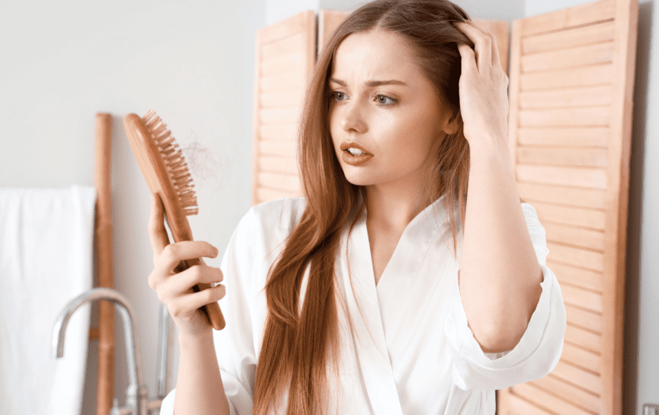 A woman with long brown hair in a white robe looks concerned as she holds a hairbrush filled with strands. Standing in her bathroom before wooden shutters, shes contemplating the best shampoos for women with thinning hair.