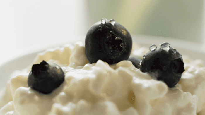 Close-up of fresh blueberries on top of creamy cottage cheese in a white bowl. The image highlights the texture and moistness of the berries and the smoothness of the cottage cheese, with a soft-focus background.