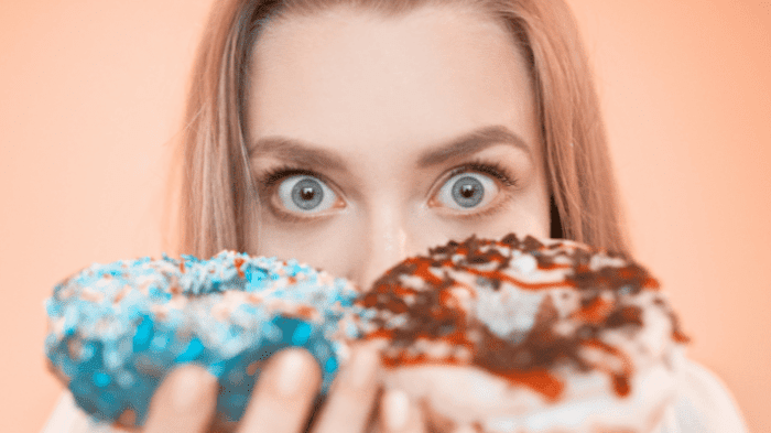 A person with wide eyes holding two doughnuts close to their face. One doughnut has blue icing with sprinkles, and the other has white icing with chocolate and red drizzle. The background is a soft peach color.