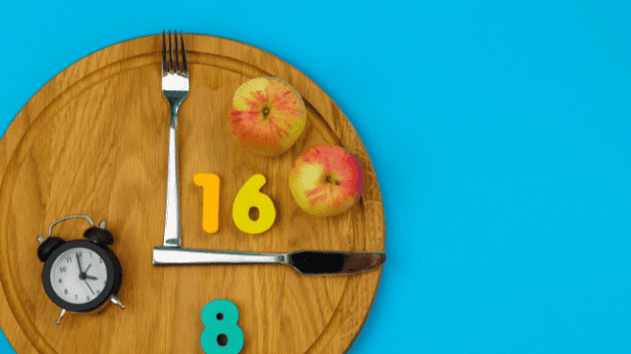 A wooden plate designed as a clock, with a fork and knife as clock hands pointing to numbers 16 and 8. Two apples are on the plate. A small black alarm clock is placed on the left. The background is bright blue.