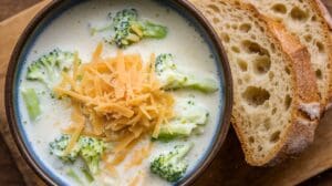 A bowl of rich broccoli cheddar soup topped with shredded cheese rests on a wooden surface, accompanied by slices of rustic bread.