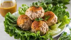 Four browned chicken meatballs garnished with fresh herbs, placed on a bed of green lettuce leaves on a white plate. Enhanced with creamy cottage cheese, a small carafe of dressing is in the background on the left.