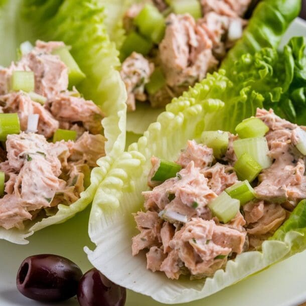 Three lettuce wraps on a white plate filled with a creamy chicken salad mixture, reminiscent of tuna salad lettuce cups, garnished with chopped celery and onions. The plate rests on a wooden board, accompanied by two black olives and cherry tomatoes in the background.