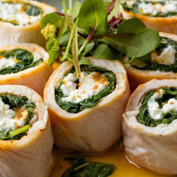 Cottage Cheese and Spinach Stuffed Chicken Roll-Ups are artfully arranged in a circle on a plate. Each rolled chicken breast is garnished with fresh greens and drizzled with a light sauce, creating an elegant and delicious presentation.