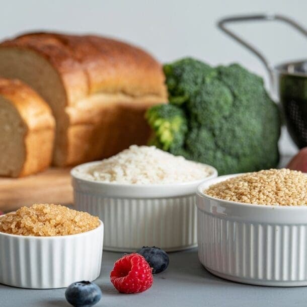 Sliced bread, broccoli, and red potatoes sit near a colander; bowls of brown sugar, white rice, and oats—examples of the best carbohydrates—are surrounded by raspberries and blueberries on a gray surface.