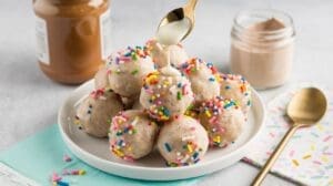 A plate of birthday cake protein balls is topped with a drizzle of white sauce from a spoon. The balls, boasting colorful sprinkles, share the scene with a jar of spread, an empty jar, a gold spoon, and folded napkins—perfect for any festive occasion.