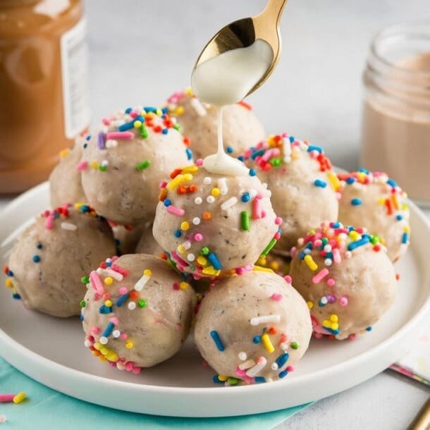 A plate of birthday cake protein balls is topped with a drizzle of white sauce from a spoon. The balls, boasting colorful sprinkles, share the scene with a jar of spread, an empty jar, a gold spoon, and folded napkins—perfect for any festive occasion.
