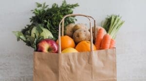 A brown paper grocery bag filled with fresh fruits and vegetables—like apples, oranges, lemons, carrots, potatoes, cabbage, and leafy greens—makes the perfect GLP-1 Shopping List against a light background.