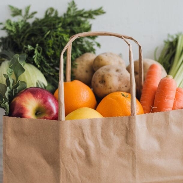A brown paper grocery bag filled with fresh fruits and vegetables—like apples, oranges, lemons, carrots, potatoes, cabbage, and leafy greens—makes the perfect GLP-1 Shopping List against a light background.