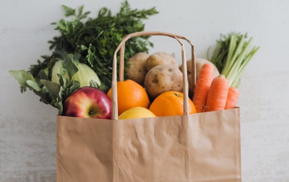 A brown paper grocery bag filled with fresh fruits and vegetables—like apples, oranges, lemons, carrots, potatoes, cabbage, and leafy greens—makes the perfect GLP-1 Shopping List against a light background.