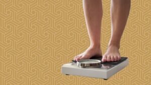 A person stands barefoot on a bathroom scale, symbolizing their fat loss journey, against a yellow geometric patterned background. The scale’s dial is visible, but the weight reading remains unclear.