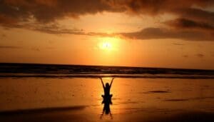 A person practices meditation for stress on a beach at sunset, arms raised and silhouetted against the orange sky reflected in wet sand. Waves roll in beneath dramatic clouds, creating a serene scene to find calm.
