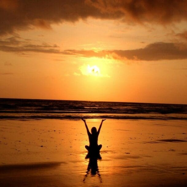 A person practices meditation for stress on a beach at sunset, arms raised and silhouetted against the orange sky reflected in wet sand. Waves roll in beneath dramatic clouds, creating a serene scene to find calm.