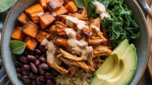 A Protein Bowl filled with BBQ chicken, creamy sauce, roasted sweet potato cubes, black beans, avocado slices, kale, and quinoa, garnished with fresh greens.