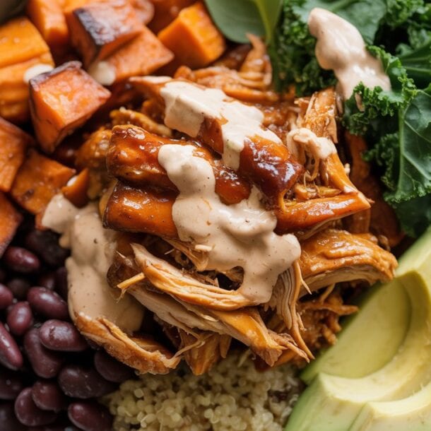 A Protein Bowl filled with BBQ chicken, creamy sauce, roasted sweet potato cubes, black beans, avocado slices, kale, and quinoa, garnished with fresh greens.