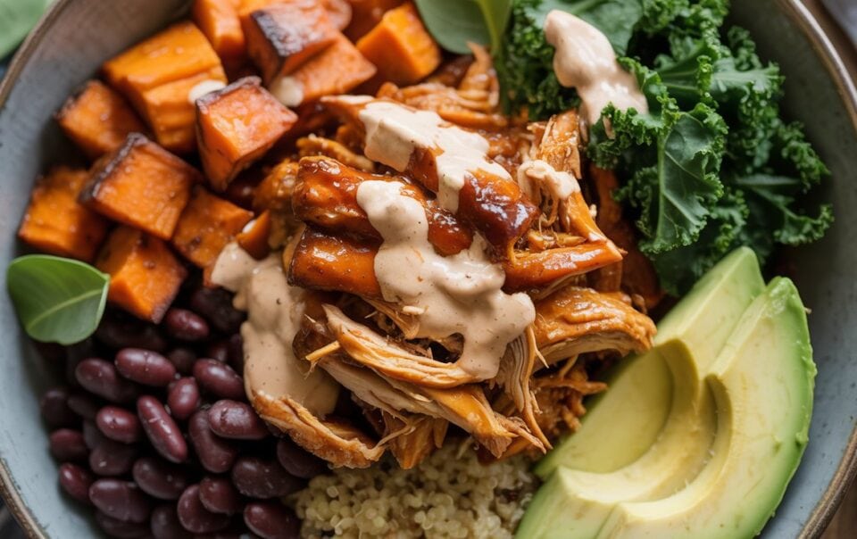 A Protein Bowl filled with BBQ chicken, creamy sauce, roasted sweet potato cubes, black beans, avocado slices, kale, and quinoa, garnished with fresh greens.