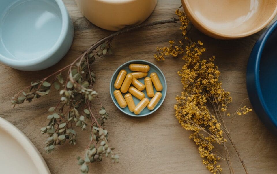 A small blue dish filled with yellow capsules sits on a wooden surface, surrounded by dried branches and bowls of various colors.