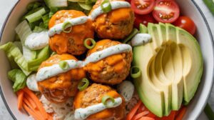 A Protein Bowl featuring rice, lettuce, shredded carrots, cherry tomato halves, avocado slices, and four saucy Buffalo Turkey Meatballs topped with creamy ranch dressing and sliced green onions.