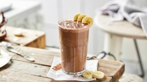 A tall glass of cottage cheese chocolate smoothie sits on a rustic wooden table, garnished with banana slices on the rim and cocoa powder sprinkled on top, with extra banana slices nearby.