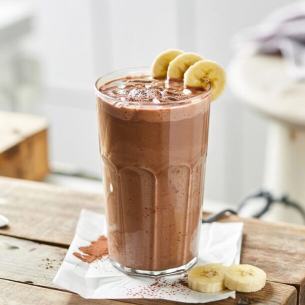 A tall glass of cottage cheese chocolate smoothie sits on a rustic wooden table, garnished with banana slices on the rim and cocoa powder sprinkled on top, with extra banana slices nearby.