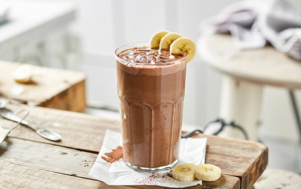 A tall glass of cottage cheese chocolate smoothie sits on a rustic wooden table, garnished with banana slices on the rim and cocoa powder sprinkled on top, with extra banana slices nearby.
