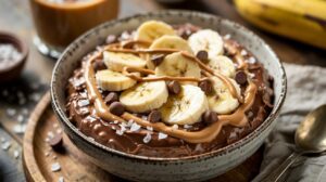 A bowl of chocolate peanut butter smoothie topped with banana slices, chocolate chips, peanut butter drizzle, and a sprinkle of sea salt, creating a delicious high-protein dessert, set on a wooden tray with a spoon and napkin nearby.