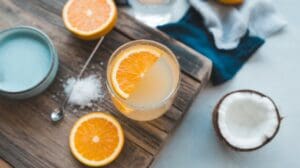 A glass of Salted OJ garnished with an orange slice sits on a wooden board, surrounded by halved oranges, a bowl of salt, a spoon, and a halved coconut. A blue cloth is partially visible in the background.