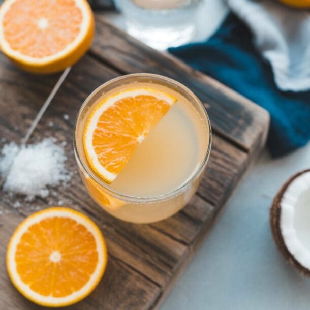 A glass of Salted OJ garnished with an orange slice sits on a wooden board, surrounded by halved oranges, a bowl of salt, a spoon, and a halved coconut. A blue cloth is partially visible in the background.