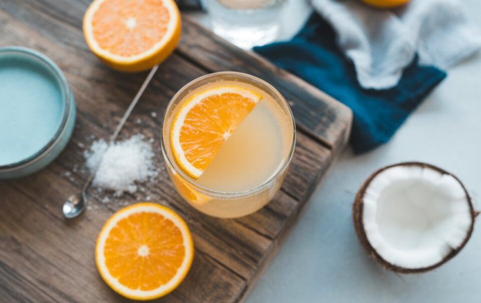 A glass of Salted OJ garnished with an orange slice sits on a wooden board, surrounded by halved oranges, a bowl of salt, a spoon, and a halved coconut. A blue cloth is partially visible in the background.