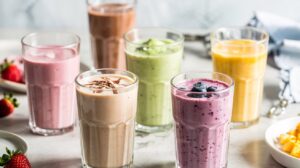 Six glasses of colorful cottage cheese smoothies—pink, yellow, green, purple, and brown—are arranged on a light surface. Some are topped with fruit, with scattered strawberries and mango pieces nearby.