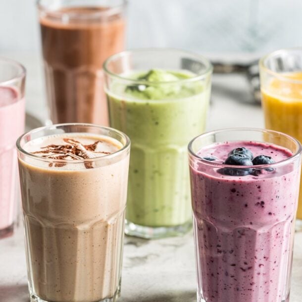 Six glasses of colorful cottage cheese smoothies—pink, yellow, green, purple, and brown—are arranged on a light surface. Some are topped with fruit, with scattered strawberries and mango pieces nearby.