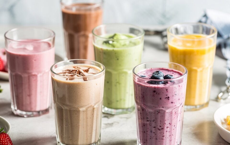 Six glasses of colorful cottage cheese smoothies—pink, yellow, green, purple, and brown—are arranged on a light surface. Some are topped with fruit, with scattered strawberries and mango pieces nearby.