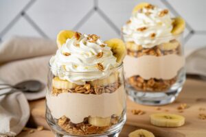 Two glass cups layered with granola, banana slices, creamy yogurt, and topped with whipped cream and granola. Sliced bananas and a gray napkin are visible on the wooden surface nearby.