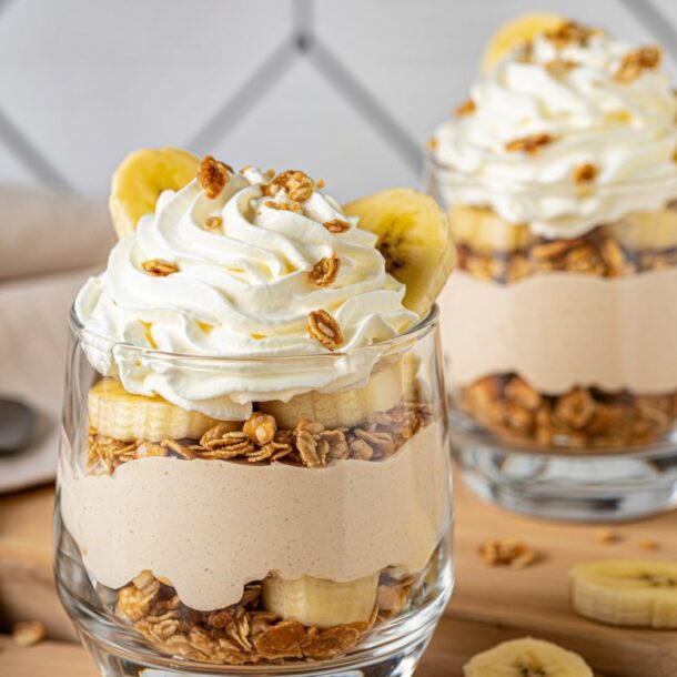 Two glass cups layered with granola, banana slices, creamy yogurt, and topped with whipped cream and granola. Sliced bananas and a gray napkin are visible on the wooden surface nearby.