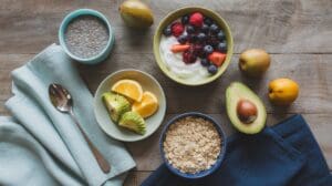 A breakfast spread with a bowl of yogurt topped with berries, a bowl of oatmeal, a bowl of chia pudding, sliced avocado, orange slices, whole avocados, kiwis, and folded napkins with a spoon on a wooden table.