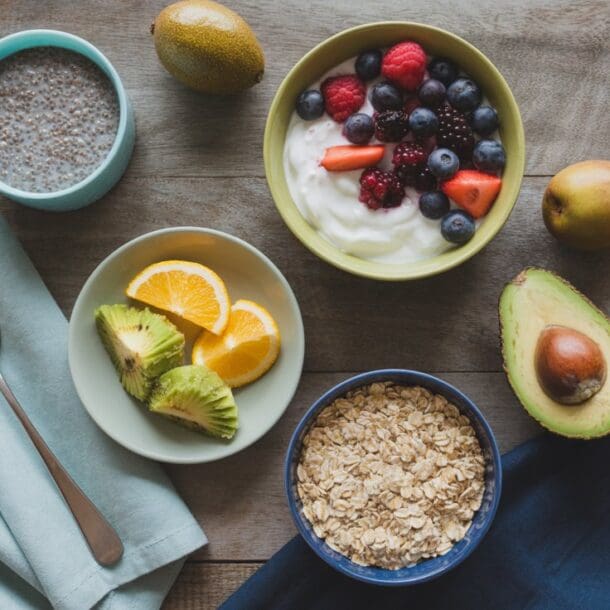 A breakfast spread with a bowl of yogurt topped with berries, a bowl of oatmeal, a bowl of chia pudding, sliced avocado, orange slices, whole avocados, kiwis, and folded napkins with a spoon on a wooden table.