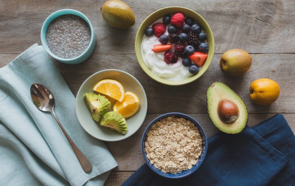 A breakfast spread with a bowl of yogurt topped with berries, a bowl of oatmeal, a bowl of chia pudding, sliced avocado, orange slices, whole avocados, kiwis, and folded napkins with a spoon on a wooden table.