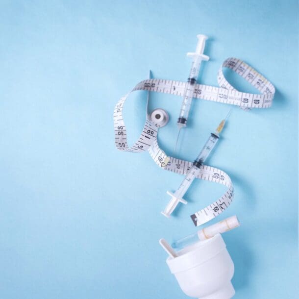 A white measuring tape is arranged in a spiral alongside syringes, a small vial, and a white container on a light blue background, symbolizing themes of medication and weight loss management.