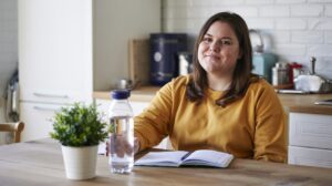 A beginner in a yellow sweater sits at a kitchen table with a notebook, bottle of water, and potted plant, smiling slightly at the camera. Kitchen appliances and utensils are visible in the background.