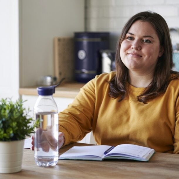 A beginner in a yellow sweater sits at a kitchen table with a notebook, bottle of water, and potted plant, smiling slightly at the camera. Kitchen appliances and utensils are visible in the background.