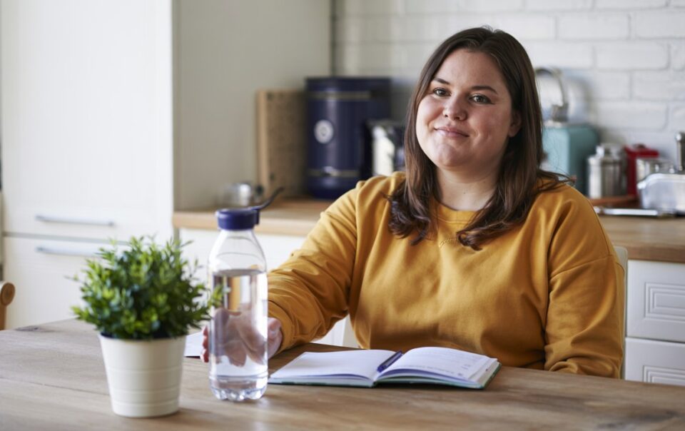 A beginner in a yellow sweater sits at a kitchen table with a notebook, bottle of water, and potted plant, smiling slightly at the camera. Kitchen appliances and utensils are visible in the background.