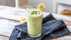 A glass of Green Cottage Cheese Smoothie, garnished with a banana slice and mint leaves, sits on a black cloth napkin atop a rustic wooden table.
