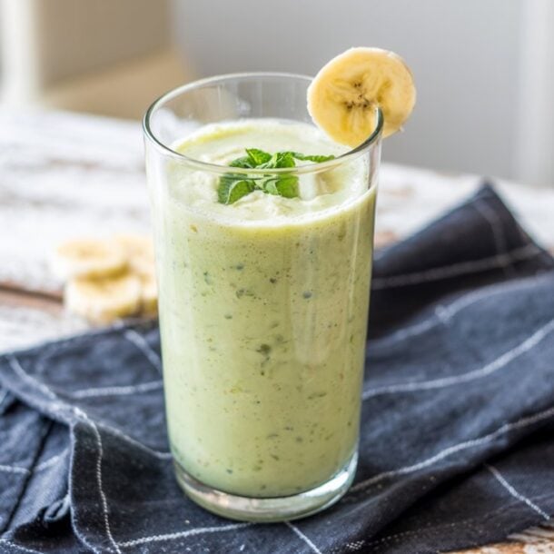 A glass of Green Cottage Cheese Smoothie, garnished with a banana slice and mint leaves, sits on a black cloth napkin atop a rustic wooden table.