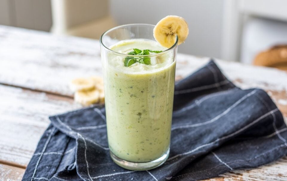 A glass of Green Cottage Cheese Smoothie, garnished with a banana slice and mint leaves, sits on a black cloth napkin atop a rustic wooden table.