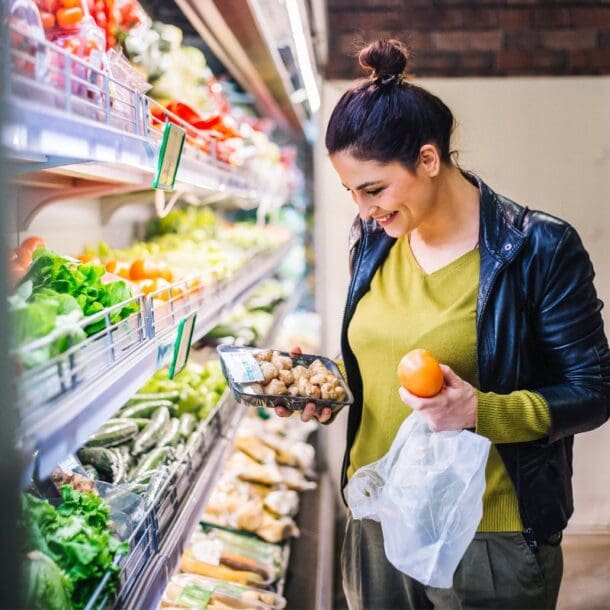 A woman smiles while grocery shopping, holding a tomato and a plastic-wrapped pack of mushrooms—perfect ingredients for GLP-1 recipes—in the produce section filled with fresh vegetables.