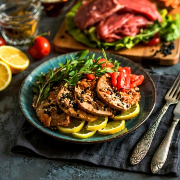 A plate of sliced cooked meat garnished with herbs, sesame seeds, and tomato pieces, served on lemon slices for added nutrition. In the background, raw meat and vegetables rest on a wooden board. Cutlery and lemon halves are nearby.