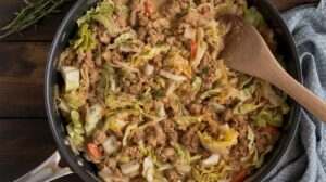 A skillet filled with cooked ground turkey, chopped cabbage, and diced vegetables, with a wooden spoon resting inside. The dish appears hearty and well-seasoned, sitting on a dark wooden surface with a cloth nearby.