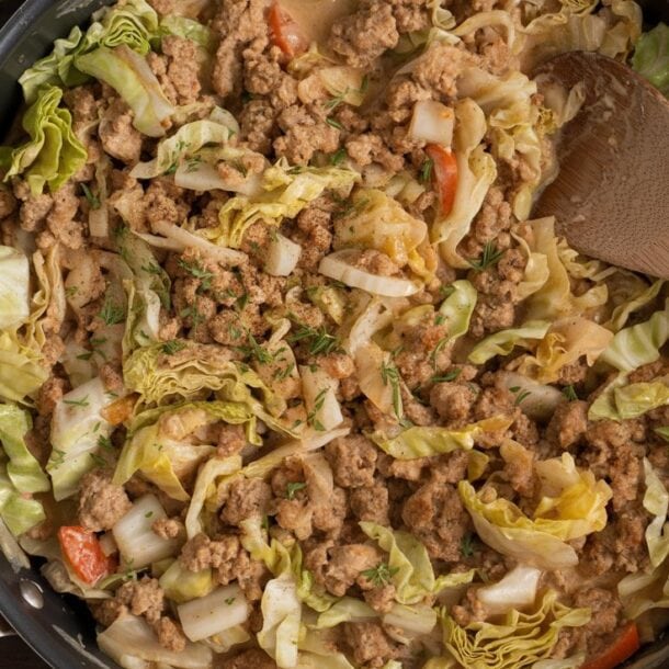 A skillet filled with cooked ground turkey, chopped cabbage, and diced vegetables, with a wooden spoon resting inside. The dish appears hearty and well-seasoned, sitting on a dark wooden surface with a cloth nearby.
